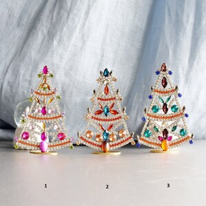 May include: Three jeweled Christmas tree ornaments, each with a different color scheme. The first is pink and white, the second is orange and white, and the third is blue and white.