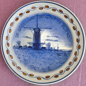 May include: A decorative ceramic plate with a blue and white windmill scene. The plate has a white border with orange leaf accents and text that reads "DE DAGERAAD BEGINTE TE BLINKEN" and "OCT. 1918."