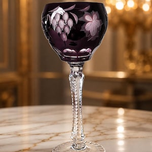 Può includere: Un calice da vino in cristallo intagliato color viola intenso con un disegno inciso di grappoli d'uva e foglie sulla coppa. Lo stelo è trasparente con un motivo a sfaccettature di diamante e la base circolare ha un motivo a raggiera.