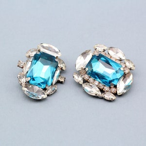 May include: A pair of blue and clear rhinestone earrings. Each earring features a large, rectangular, blue rhinestone surrounded by smaller, clear rhinestones.