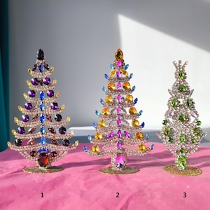 May include: Three decorative Christmas trees made of sparkling crystals. The trees are in different colors: purple, blue, yellow, and green. Each tree has a unique design and sits on a gold-colored base. The trees are numbered 1, 2, and 3.