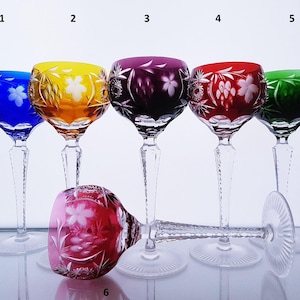 May include: Six cut-to-clear crystal wine glasses in blue, yellow, purple, red, green, and pink. Each glass features intricate floral and star-shaped engravings on the bowl, a faceted clear stem, and a circular base. Numbers 1 through 6 are visible.