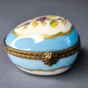 May include: An egg-shaped porcelain trinket box featuring a glossy light blue base and a white top decorated with hand-painted purple flowers and green leaves. The two halves are joined by a decorative brass hinge with a shell-shaped clasp.