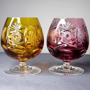 May include: Two cut crystal brandy glasses, one in amber and one in purple, featuring intricate starburst and geometric patterns. Each glass has a wide, rounded bowl, a short stem, and a clear, circular base with a radial, sunburst-like design.