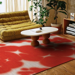 May include: A red and white patterned rug with a floral design. The rug is placed on a wooden floor, with a yellow sofa and a white oval coffee table with wooden legs in the background. The room is decorated with plants.