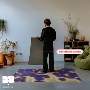 Person holding and showing the back of the rug with a slip-resistant backing showcase in a living room, highlighting a demonstration of the vibrant purple, orange, yellow and green hues animal print with faux textile and weaving hand painted texture, a printed area rug for the maximalist eclectic home.