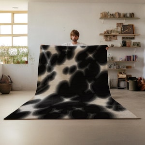 May include: A large, rectangular rug with a black and cream abstract pattern. The rug features a repeating design of organic shapes and is displayed in a well-lit room. The rug's design adds a modern touch to the space.