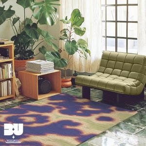 An eclectic abstract cowhide floral area rug is placed in a cool and cozy apartment, featuring books in cabinets, large houseplants, and sage green single sofa on green tiled floor. This rug showcases hand painted cowhide and floral patterns, embodying an aura and psychedelic style with vivid hues of purple, yellow, orange, and green. It’s perfect for dopamine home decor and aesthetic decor lovers.