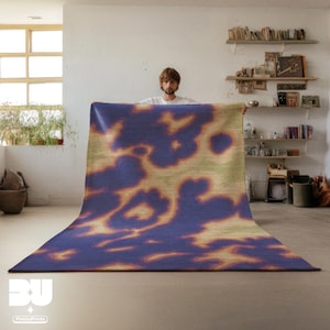 Person holding a dopamine decor, Purple Aura Psychedelic Area Rug, with abstract cowhide, animal print pattern, with vivd green and yellow hues, standing in a cozy room with books and decor on shelves.