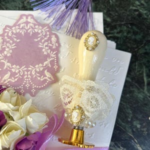 May include: A white and gold wax seal stamp with a cameo design and a lace trim. The stamp is on a white surface with a purple ribbon and a bouquet of purple and white flowers.