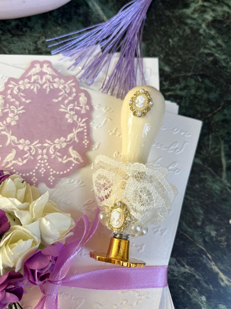 May include: A white and gold wax seal stamp with a cameo design and a lace trim. The stamp is on a white surface with a purple ribbon and a bouquet of purple and white flowers.