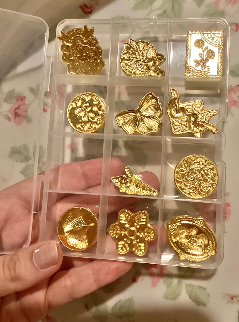 May include: A clear plastic storage box with 12 compartments, each containing a gold-colored metal charm. The charms feature various designs, including a butterfly, a snowflake, a ginkgo leaf, and a seashell.
