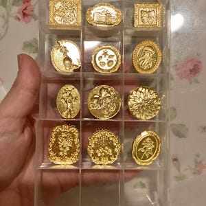 May include: A clear plastic storage case with 12 gold-colored metal stamp designs. The stamps feature various floral, geometric, and abstract designs.