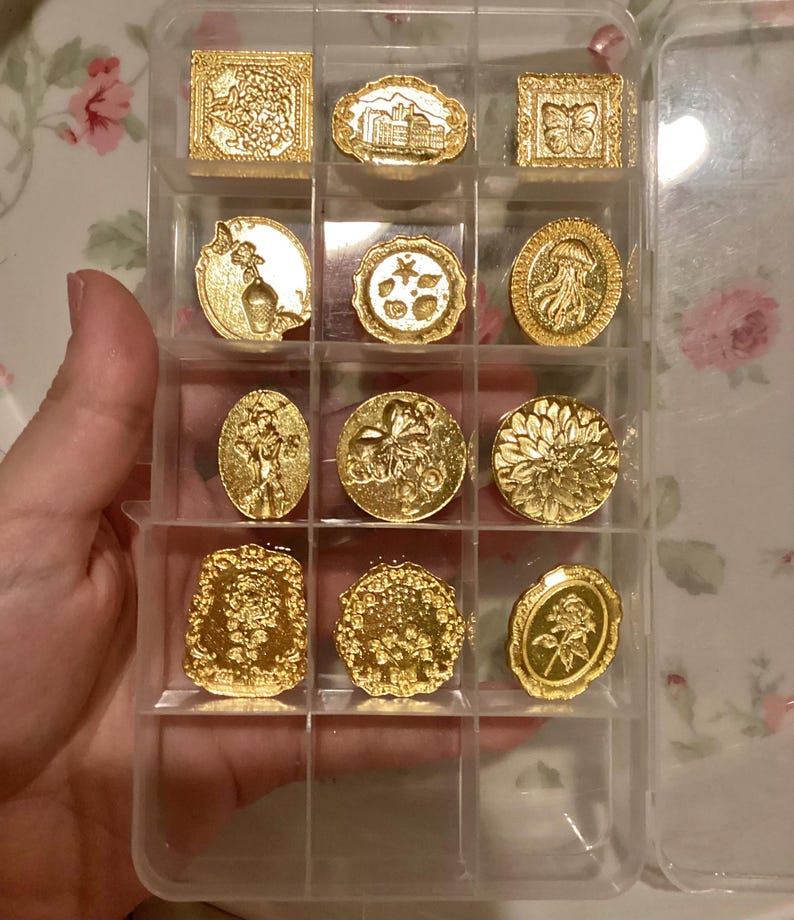 May include: A clear plastic storage case with 12 gold-colored metal stamp designs. The stamps feature various floral, geometric, and abstract designs.