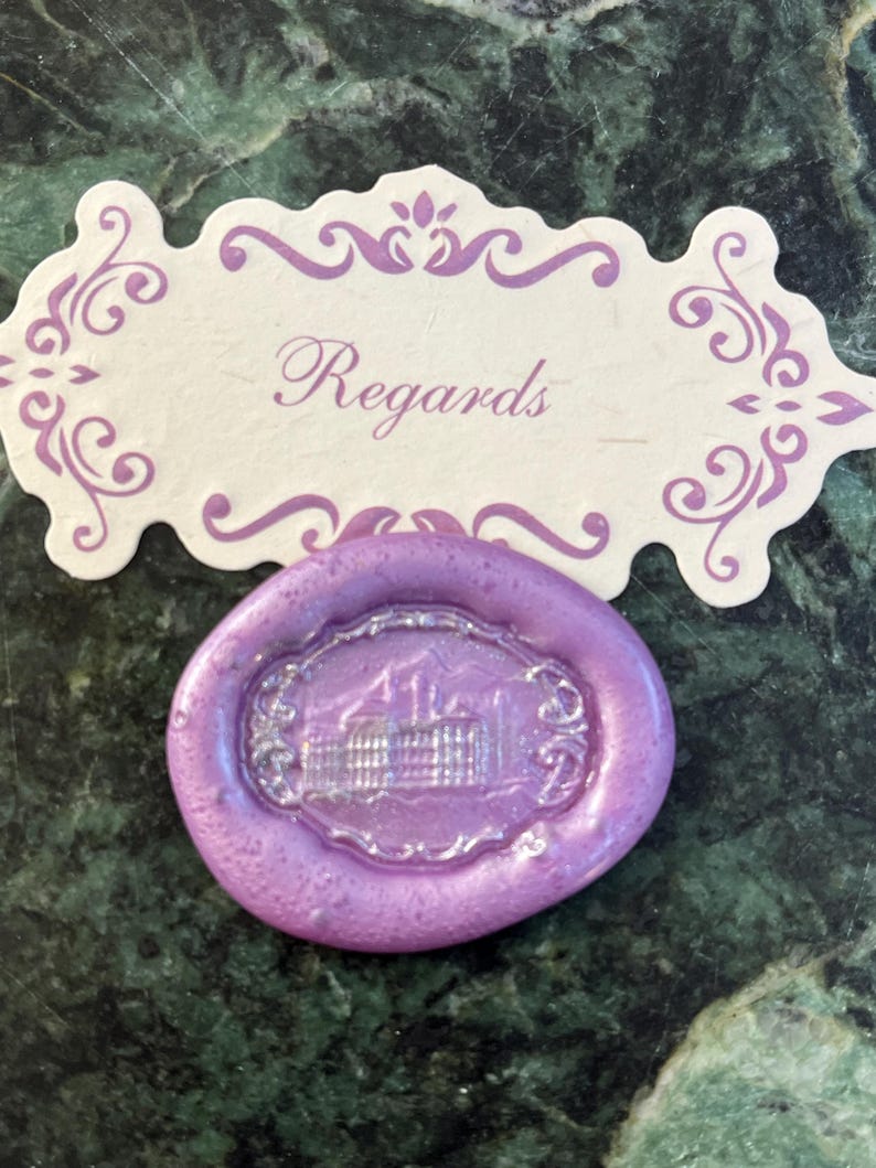May include: A purple wax seal with a silver embossed design of a building. The seal is on a white card with the word "Regards" written in purple.