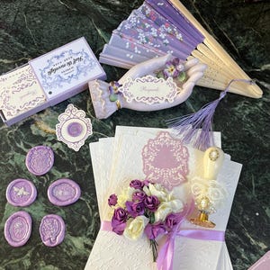 May include: A set of purple wax seals, a box of notecards with the text "Just Say Bonjour" on the front, a porcelain hand holding a card that says "Regards", a purple and white fan, and a stack of white envelopes with a purple floral design.