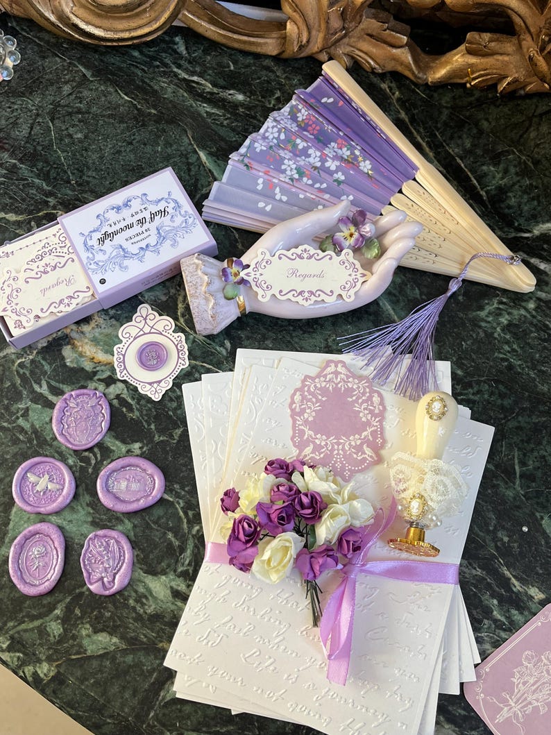 May include: A set of purple wax seals, a box of notecards with the text "Just Say Bonjour" on the front, a porcelain hand holding a card that says "Regards", a purple and white fan, and a stack of white envelopes with a purple floral design.
