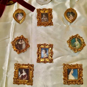 Könnte beinhalten: Eine Sammlung kleiner, verzierter goldener Bilderrahmen, die jeweils ein Miniaturporträt einer Frau enthalten. Die Rahmen, in verschiedenen Formen wie Herz und Quadrat, sind auf einer weißen, strukturierten Oberfläche angeordnet. Eine weiße Rose und eine dekorative Hand sind ebenfalls vorhanden.