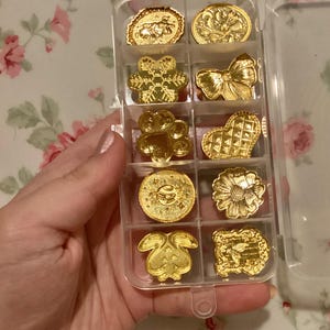 May include: A clear plastic storage box with 10 gold-colored metal charms. The charms are in the shape of a snowflake, paw print, heart, flower, bunny, bow, and other designs.