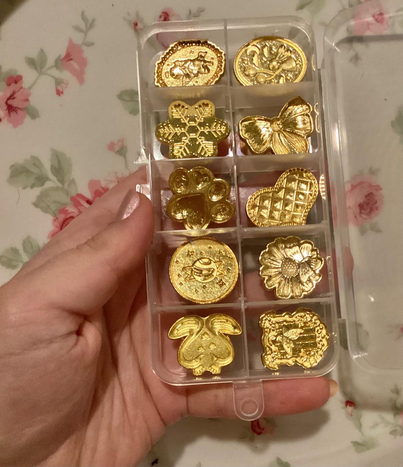 May include: A clear plastic storage box with 10 gold-colored metal charms. The charms are in the shape of a snowflake, paw print, heart, flower, bunny, bow, and other designs.