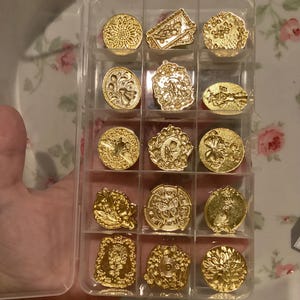 May include: A clear plastic storage case filled with 15 gold-toned metal stamp designs. The stamps feature various floral and geometric patterns.