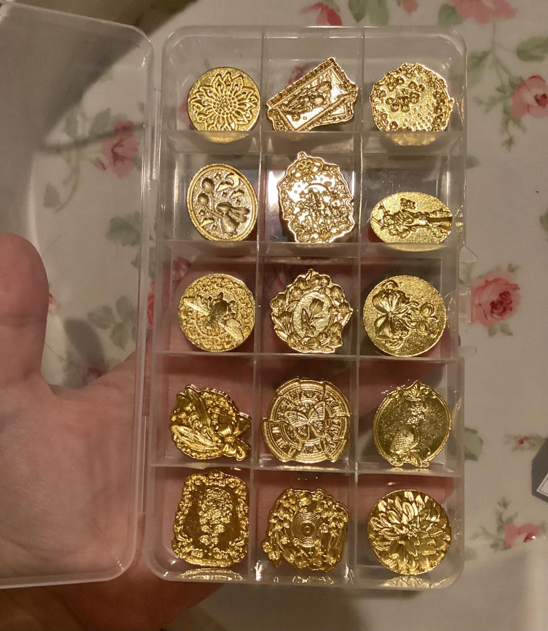 May include: A clear plastic storage case filled with 15 gold-toned metal stamp designs. The stamps feature various floral and geometric patterns.