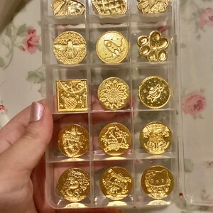 May include: A clear plastic storage case with 15 gold-colored metal stamps. The stamps feature various designs, including a heart, a sunflower, a rocket ship, a bee, a paw print, and a cat.
