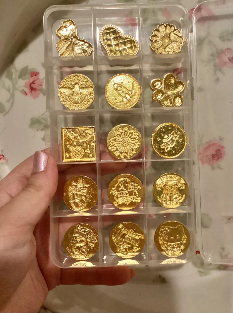May include: A clear plastic storage case with 15 gold-colored metal stamps. The stamps feature various designs, including a heart, a sunflower, a rocket ship, a bee, a paw print, and a cat.