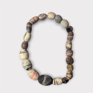Puede incluir: Un collar hecho de cuentas de lana afieltrada en varios tonos de gris, marrón y crema. Las cuentas ovaladas tienen rayas y patrones en naranja, negro y rosa. El collar se muestra sobre un fondo blanco.