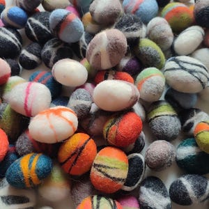 Könnte beinhalten: Nahaufnahme einer Ansammlung von Filzkugeln aus Wolle in verschiedenen Farben und Mustern. Die Kugeln sind mit Streifen und Designs in Blau-, Orange-, Rot-, Grün-, Weiß- und Grautönen verziert. Ideal für Bastelarbeiten und Schmuckherstellung.