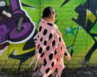 May include: A pink shawl with large black cut-out shapes and fringed edges. The shawl is draped over the shoulders. The background features a green brick wall with graffiti art.