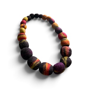 Puede incluir: Un collar compuesto por cuentas de fieltro esféricas de diferentes tamaños. Las cuentas presentan un color base oscuro con acentos en espiral de rojo, naranja, amarillo y morado. Las cuentas están dispuestas en un patrón circular continuo.