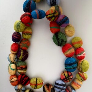Puede incluir: Un collar colorido hecho de cuentas de lana afieltrada. Las cuentas ovaladas presentan una variedad de colores, incluyendo rojo, azul, verde, amarillo y morado. Cada cuenta tiene un patrón de rayas único. El collar está ensartado en un cordón transparente.
