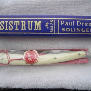 Puede incluir: Una navaja de afeitar recta vintage con mango blanco y una tapa de metal dorada. La navaja está en una caja azul con letras doradas que dicen "SISTRUM" y "Paul Drees Solingen, Alemania".