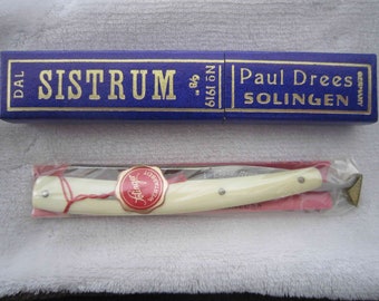 Puede incluir: Una navaja de afeitar recta vintage con mango blanco y una tapa de metal dorada. La navaja está en una caja azul con letras doradas que dicen "SISTRUM" y "Paul Drees Solingen, Alemania".