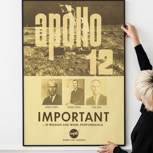 Op de afbeelding: Een vintage poster met een zwarte lijst met een foto van het maanoppervlak met de tekst "Apollo 12" in grote, vetgedrukte, gele letters. Onder de foto staan drie kleinere foto's van de astronauten Charles Conrad, Richard Gordon en Allen Bean. De poster bevat ook de tekst "IMPORTANT... IN MISSION AND WORK PERFORMANCE" en het NASA-logo met de tekst "MANNED FLIGHT AWARENESS".