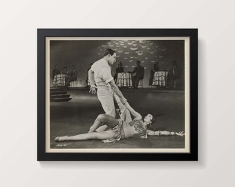 May include: Black and white photograph of a man and woman dancing. The man is wearing a white shirt and the woman is wearing a dark dress. They are dancing in a nightclub setting with tables and chairs in the background.