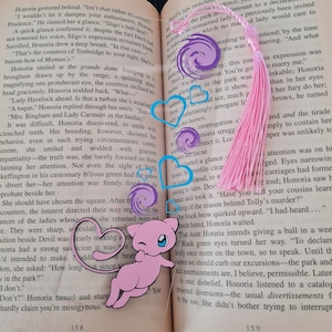 May include: A pink and purple bookmark with a pink tassel. The bookmark features a cartoon image of a pink Pokemon character with a winking eye. The bookmark is placed on an open book with text about a social gathering.