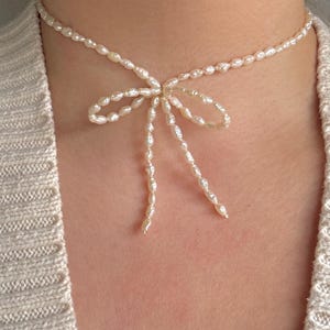 May include: A delicate pearl choker necklace with a bow made of pearls. The necklace is white and the pearls are small and irregular in shape.