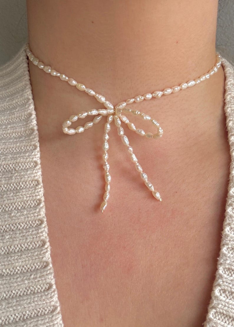 May include: A delicate pearl choker necklace with a bow made of pearls. The necklace is white and the pearls are small and irregular in shape.