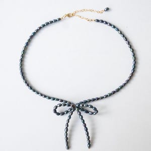 May include: A delicate necklace made of dark gray pearls with a bow tied at the center. The necklace is made of small, oval-shaped pearls.