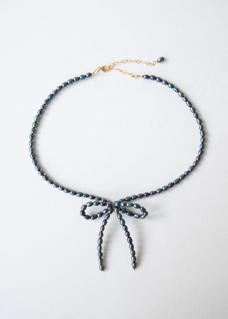 May include: A delicate necklace made of dark gray pearls with a bow tied at the center. The necklace is made of small, oval-shaped pearls.