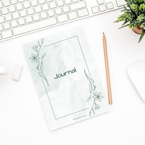 May include: A green and white journal with a floral design and the word "Journal" in a black frame. The journal is on a white surface with a pencil and a keyboard.