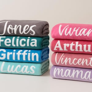 K&ouml;nnte beinhalten: Ein Stapel personalisierter Decken in verschiedenen Farben, darunter Grau, T&uuml;rkis, Blau und Rosa. Jede Decke ist mit einem Namen in wei&szlig;er Schrift bestickt. Die Namen sind "Jones", "Felicia", "Griffin", "Lucas", "Vivian", "Arthur", "Vincent" und "mama".