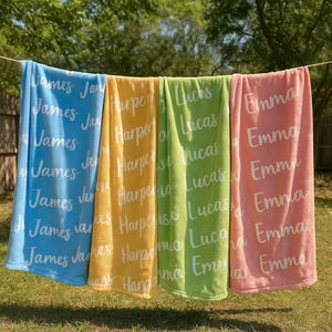 K&ouml;nnte beinhalten: Vier personalisierte Strandt&uuml;cher h&auml;ngen an einer W&auml;scheleine. Die Handt&uuml;cher sind in Blau, Gelb, Gr&uuml;n und Rosa gehalten. Jedes Handtuch hat einen Namen in wei&szlig;er Schrift: James, Harper, Lucas und Emma.