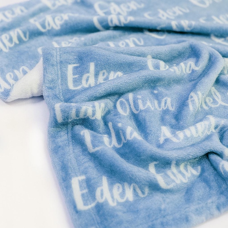 K&ouml;nnte beinhalten: Eine hellblaue Decke mit wei&szlig;en Schriftz&uuml;gen. Die Namen "Eden", "Camden", "Lilia" und "Olivia" sind zu sehen. Die Decke hat eine weiche Textur und eine wei&szlig;e R&uuml;ckseite.