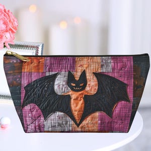 May include: A purple and orange patchwork cosmetic bag with a black bat design. The bat has a yellow face and orange eyes.