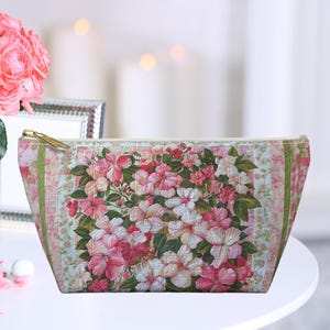 May include: A floral print cosmetic bag with a light green background and pink flowers. The bag has a gold zipper and is sitting on a white table.