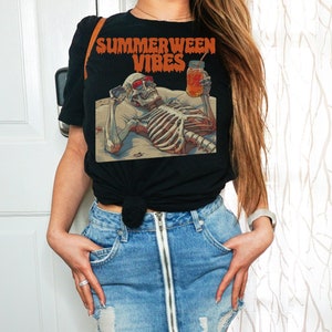 May include: A black t-shirt with the text "Summerween Vibes" and a skeleton graphic holding a glass of orange juice. The person is wearing a blue denim skirt with a zipper down the front.