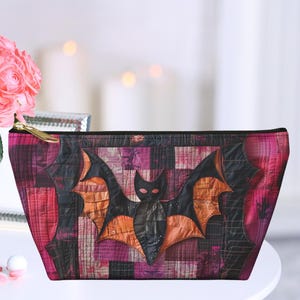 May include: A pink and black quilted makeup bag featuring a bat design. The bat is made from patchwork fabric and has a black face with white eyes.